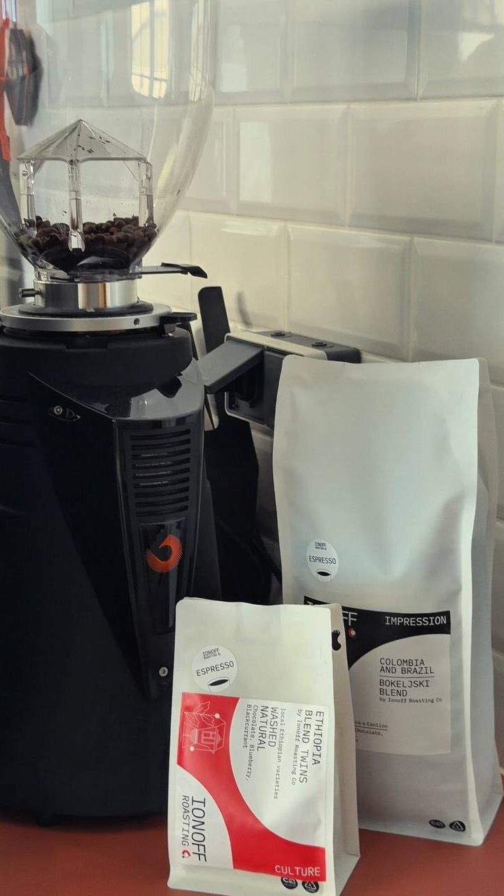 Two bags of Ionoff Roasting Co. coffee next to the grinder, in front of white tiles.